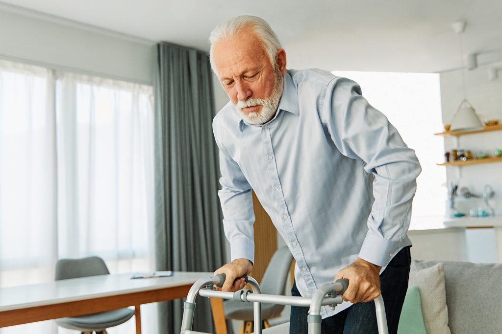 Elderly man with white hair and beard using a walker in a bright living room. He wears a light blue shirt and stands near a wooden table with chairs. Large windows and curtains are in the background, along with shelves holding books and decor.