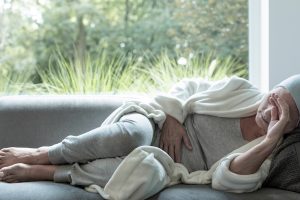 A person in a gray outfit and white robe is lying on a gray sofa, with one arm covering their eyes. They appear to be resting or experiencing discomfort. The background shows a large window with a view of green trees and plants, indicating a bright and serene outdoor setting.