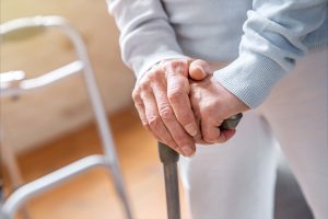 An elderly person wearing a light blue sweater and white pants grips the handle of a cane with both hands. A metal walker is visible in the blurred background. The setting appears to be indoors, with warm lighting.