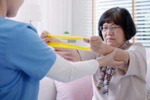 An elderly woman in glasses performs a resistance band exercise, assisted by a healthcare professional in blue scrubs. They are in a bright room with a white couch and a window with blinds in the background. The woman appears focused, and a pink cushion is visible beside her.