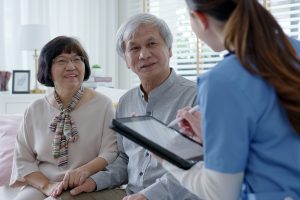 A senior Asian couple sits on a sofa, smiling, while a nurse in blue scrubs, holding a digital tablet, engages with them. The couple consists of a woman in a blouse with a colorful scarf and a man in a gray shirt. The setting is a bright, modern living room with blinds partially open.